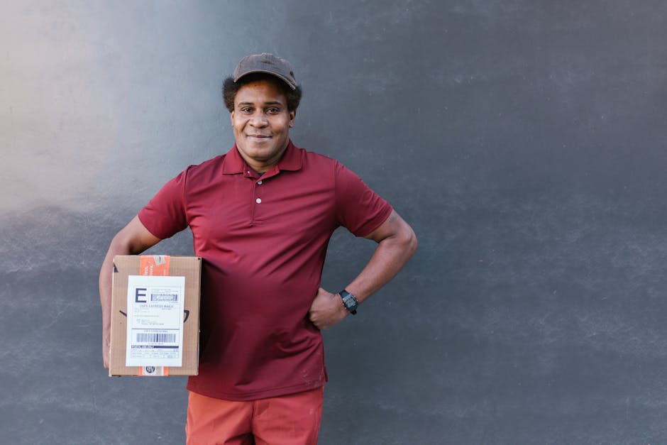 A smiling male delivery person wearing a maroon polo shirt and a grey cap, standing outdoors against a plain dark grey wall. He holds a medium-sized cardboard box with visible shipping labels, positio
