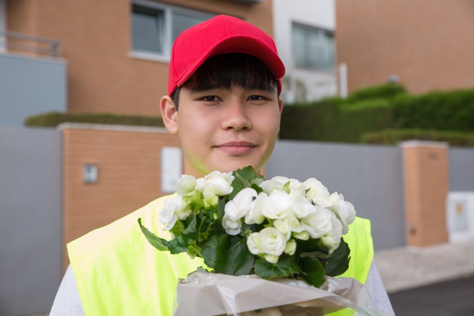 A young male flower delivery person wearing a bright yellow vest and a red cap holds a bouquet of fresh white flowers, including roses and lisianthus, wrapped in light brown paper. He stands outdoors 