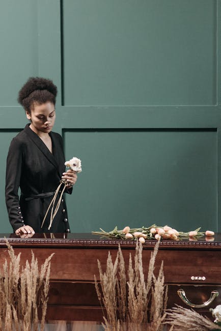 A young woman with dark curly hair styled in a natural updo, dressed in a black blazer, stands beside a wooden casket during a funeral service, holding a single white flower with a stem and a ribbon. 