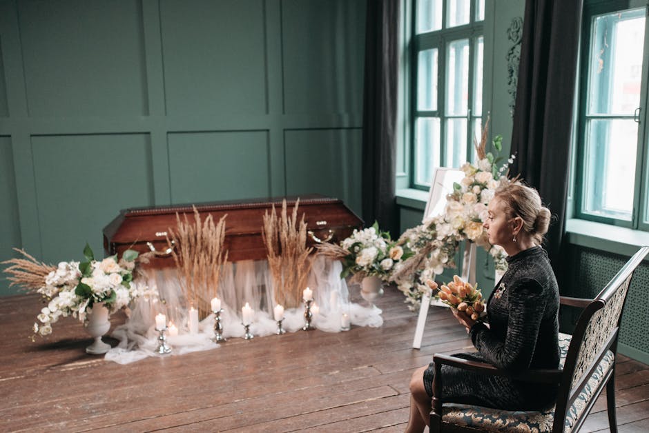 A woman seated in a patterned chair holds a bouquet of peach and cream-colored flowers, dressed in a dark textured outfit. In front of her, a floral arrangement featuring white roses, pale pink blooms