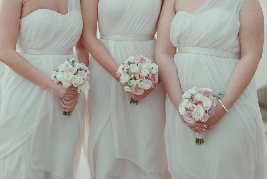 Close-up of three bridesmaids holding round bouquets of fresh flowers, featuring light pink and white roses, cream-colored ranunculus, and green foliage, wrapped with soft pastel pink and white ribbon
