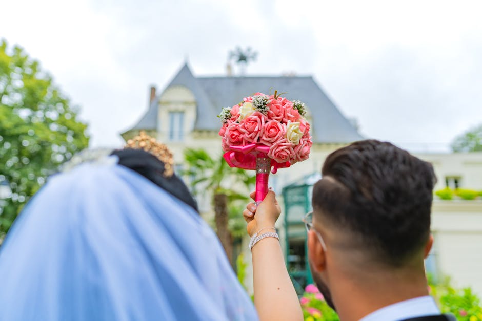 A person holding a wedding bouquet composed of pink and cream roses, with small white filler flowers, wrapped in pink ribbon, positioned outdoors in front of a historic house with grey slate roof and 