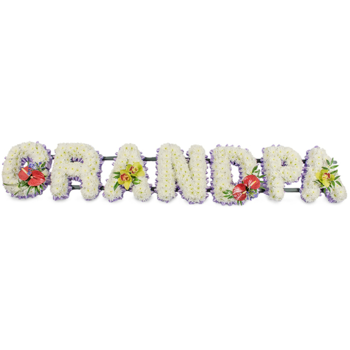 This image shows a striking sympathy flower tribute that spells the word GRANDPA in bold, three-dimensional block capitals, designed to lie horizontally as a single, continuous piece. Behind the tribute is a plain white background, which allows every flower and ribbon detail to stand out clearly. Each letter is completely blanketed in tiny white chrysanthemums, or similar small-headed blooms, set so close together that no gaps appear; the surface looks like a soft, cloud-like cushion, suggesting comfort, serenity, and purity of remembrance. Around the base of every letter runs a narrow band of pale lavender ribbon, catching the light in places and adding a graceful lilac halo beneath the fresh white petals. This pastel edging brings a gentle, dignified colour accent that feels appropriate for a funeral service or celebration of life in the Denmark Hill area. To avoid the design feeling too stark, the florist has added rich accent sprays. On the far-left G, a cluster of deep scarlet anthuriums with their distinctive heart-shaped, high-gloss petals and upright yellow spadices curves to one side, supported by dark green, tropical-style foliage. The middle D mirrors this with a similarly bold grouping of red anthuriums, creating a pleasing balance across the tribute. On the first A and the last A, arching stems of yellow-green orchids, their petals slightly translucent and delicately freckled, rest on a cushion of greenery, introducing a sense of elegance and enduring beauty. Crafted with the care you would expect from an experienced local florist, this GRANDPA tribute is the kind of arrangement families in Denmark Hill might choose for same-day delivery to a funeral home or cemetery, offering a personalised and deeply respectful farewell to a beloved grandfather.