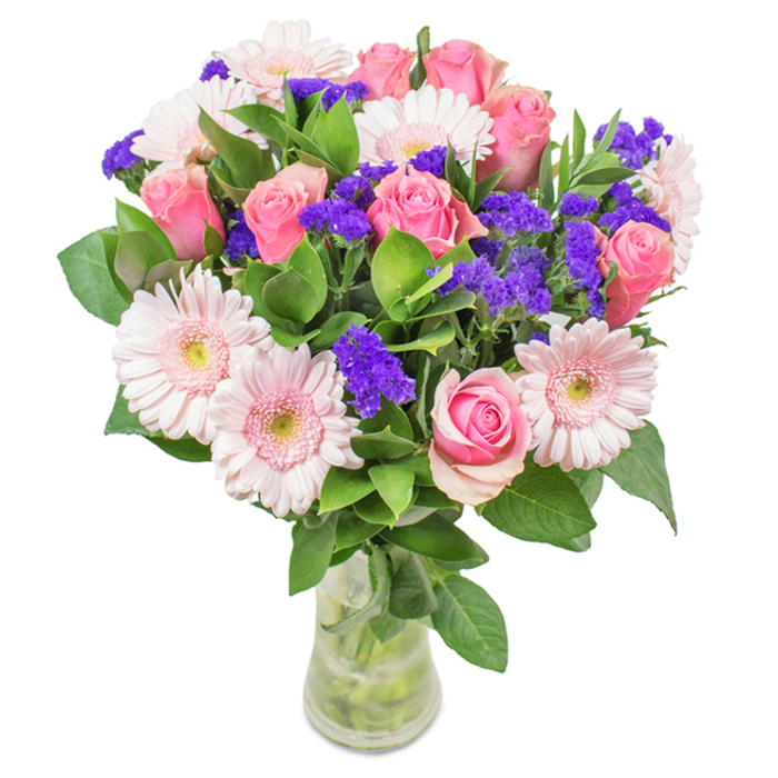 A bright, elegant floral display captured in clear glass, this bouquet is a celebration of colour and local artistry, set against a crisp white backdrop like the freshly painted shopfronts along Denmark Hill. The composition features approximately seven to nine medium-sized pink roses-some in the early, tightly wrapped stages and others just beginning to open, revealing their classic spiral forms. Encircling the roses are five to seven pale pink gerbera daisies, their generous round faces and lightly ruffled petals lending airy softness, with nearly white centres that seem to glow from within. Intertwined between the larger blooms are vivid purple accents, likely statice or limonium, adding textural intrigue and a pleasing pop of rich colour that stands out much like the community mosaics in Camberwell Green. The base of the bouquet is woven with abundant deep green foliage, enhancing freshness and vibrancy, and filling the clear vase with life and movement. Visible stems and fresh water amplify the impression of just-cut quality, reminiscent of a bespoke bouquet delivered straight to a home in Denmark Hill or nearby Herne Hill. This arrangement-combining pink roses, gerberas, and purple filler-offers a joyful note for anniversaries, birthdays, or as a distinguished centrepiece, imparting feelings of celebration, gratitude, and the warm welcome characteristic of our neighbourhood.